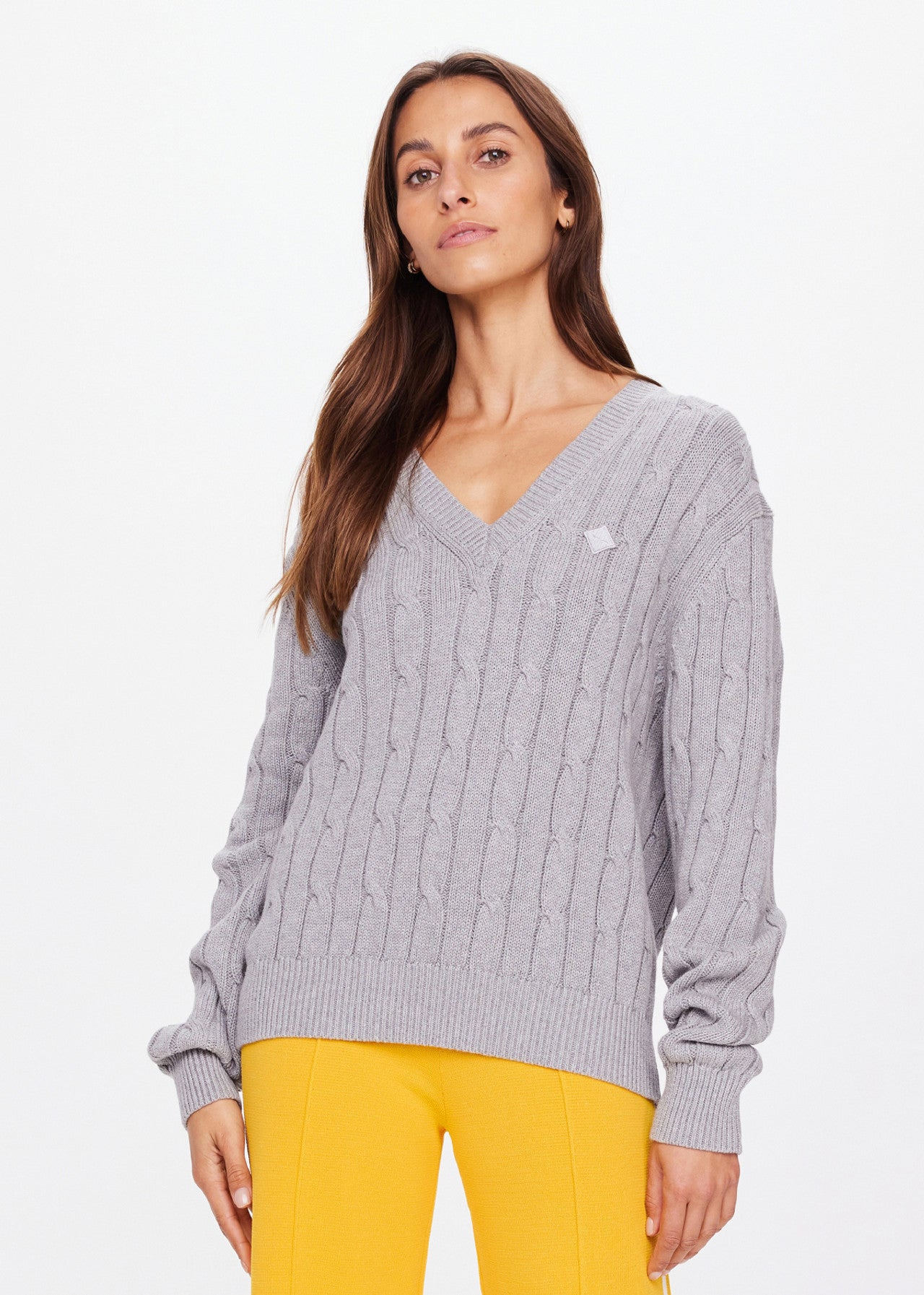 Wearing The Upside's SEYMOUR CABLE KNIT SWEATER in gray, crafted from organic cotton, exudes casual luxury when paired with vibrant yellow pants against a plain white background.