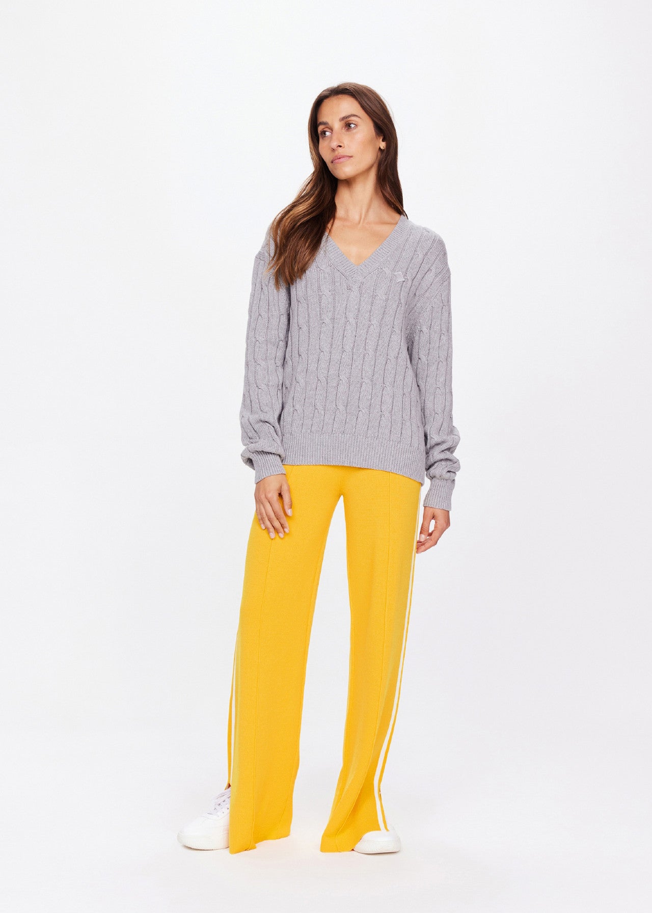 A woman models The Upside's SEYMOUR CABLE KNIT SWEATER in gray organic cotton, paired with bright yellow flared pants featuring white side stripes. With long brown hair, she gazes sideways and completes the outfit with white sneakers, embodying casual luxury against a plain white backdrop.