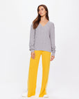 A woman models The Upside's SEYMOUR CABLE KNIT SWEATER in gray organic cotton, paired with bright yellow flared pants featuring white side stripes. With long brown hair, she gazes sideways and completes the outfit with white sneakers, embodying casual luxury against a plain white backdrop.