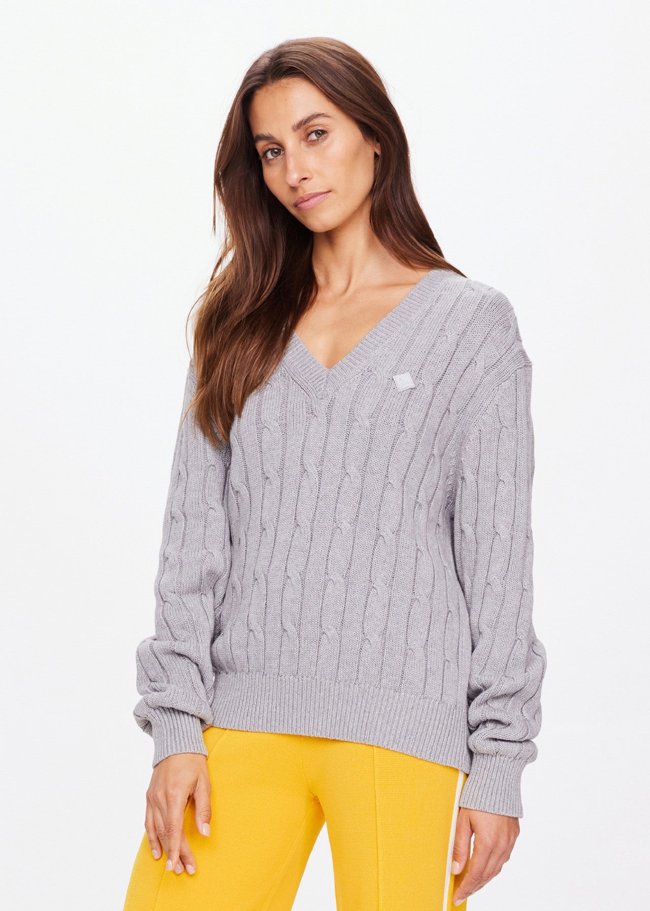 Standing against a plain white background, a person with long brown hair wears The Upside's SEYMOUR CABLE KNIT SWEATER, paired with yellow pants, embodying casual luxury.