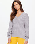 Standing against a plain white background, a person with long brown hair wears The Upside's SEYMOUR CABLE KNIT SWEATER, paired with yellow pants, embodying casual luxury.