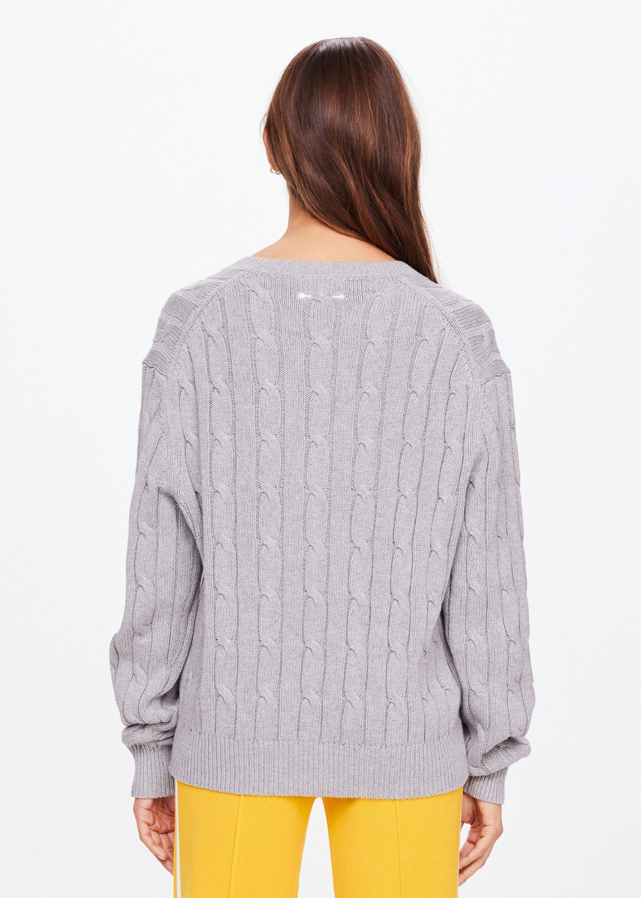 A person with long brown hair, facing away, is wearing The Upside's SEYMOUR CABLE KNIT SWEATER in elegant gray organic cotton and bright yellow pants, exuding casual luxury against a plain white background.