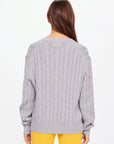 A person with long brown hair, facing away, is wearing The Upside's SEYMOUR CABLE KNIT SWEATER in elegant gray organic cotton and bright yellow pants, exuding casual luxury against a plain white background.