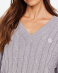 Close-up of a person with long brown hair wearing The Upside's SEYMOUR CABLE KNIT SWEATER in light gray. This V-neck sweater, made from organic cotton, features a small square logo on the left chest, exuding casual luxury.