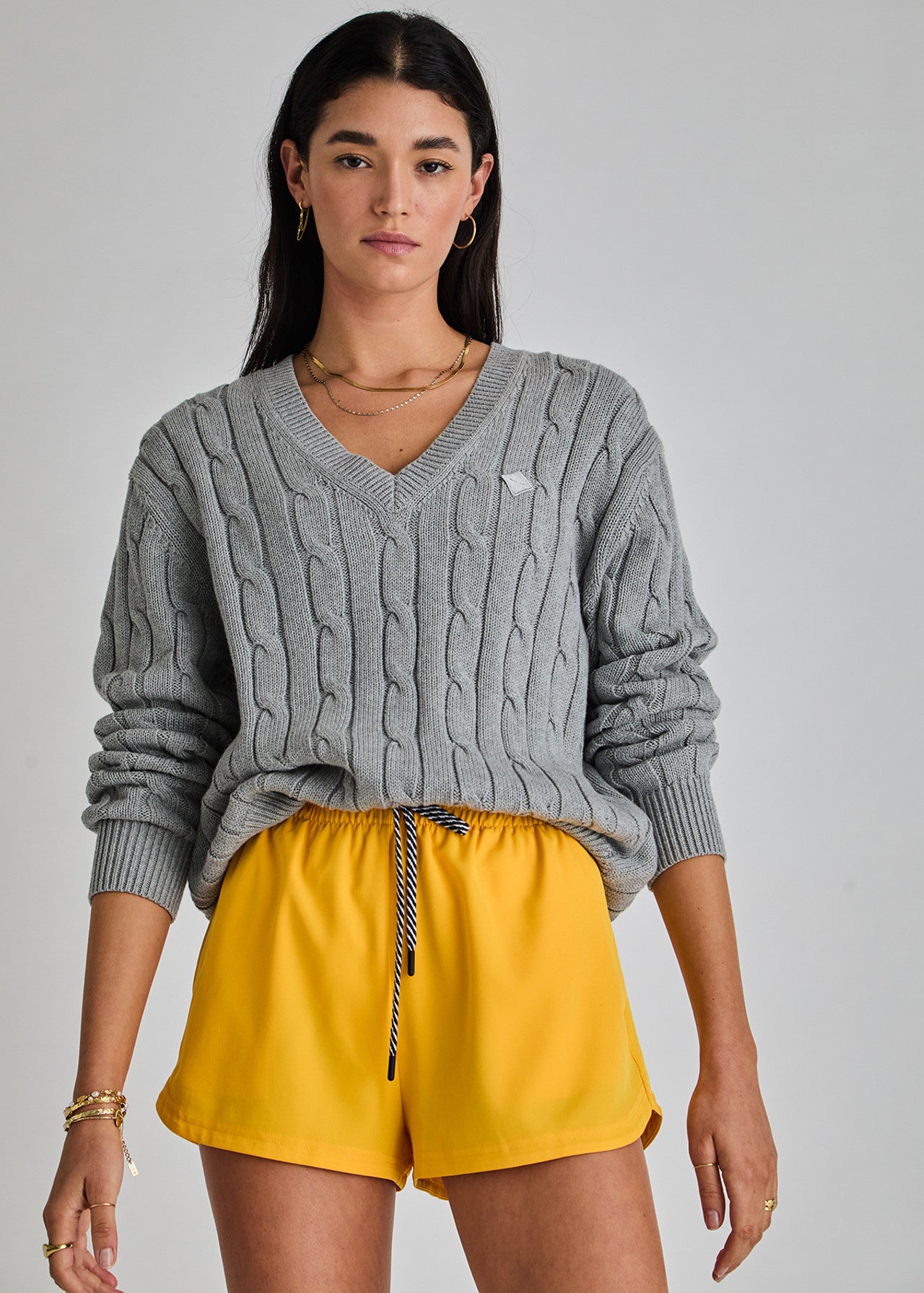 A person with long dark hair stands against a plain background in The Upside's SEYMOUR CABLE KNIT SWEATER, embodying casual luxury. They wear organic cotton yellow shorts, gold hoop earrings, and multiple bracelets.