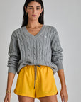 A person with long dark hair stands against a plain background in The Upside's SEYMOUR CABLE KNIT SWEATER, embodying casual luxury. They wear organic cotton yellow shorts, gold hoop earrings, and multiple bracelets.