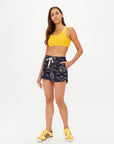 A woman with long brown hair stands against a white background wearing a yellow sports bra, The Upside Gulf Billie Run Short in navy Batik print made from recycled polyester, and yellow Adidas sneakers, with one hand in her pocket.