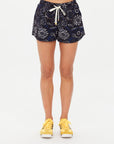 A person wearing The Upside Gulf Billie Run Short in navy blue with a white Batik print and white drawstring, paired with yellow sneakers, made from recycled polyester, stands against a plain white background.