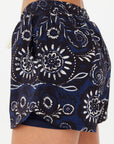 A close-up side view shows a person wearing The Upside Gulf Billie Run Short in navy blue and black with a white and blue Batik print, featuring an elastic waistband, curved hem, and crafted from recycled polyester.