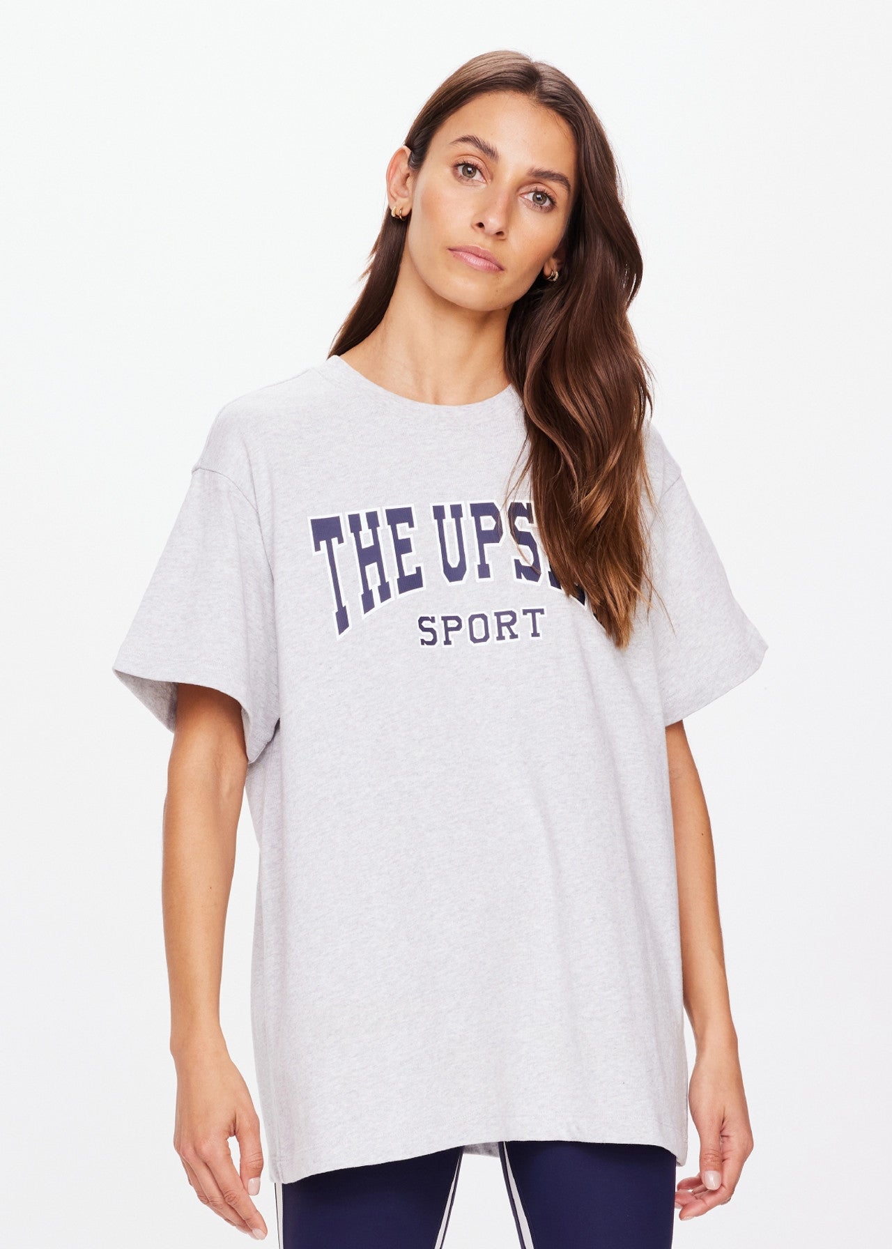 A person with long brown hair wears an oversized, organic cotton HARBOR SAMMI TEE by The Upside, featuring a navy white "THE UPSIDE SPORT" logo. They stand against a plain white background, blending comfort and style effortlessly.