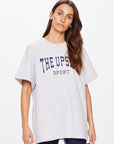 A person with long brown hair wears an oversized, organic cotton HARBOR SAMMI TEE by The Upside, featuring a navy white "THE UPSIDE SPORT" logo. They stand against a plain white background, blending comfort and style effortlessly.