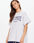 A person with long brown hair wears an oversized HARBOR SAMMI TEE made from organic cotton by The Upside, featuring a navy white logo "THE UPSIDE SPORT." They stand against a plain white background.