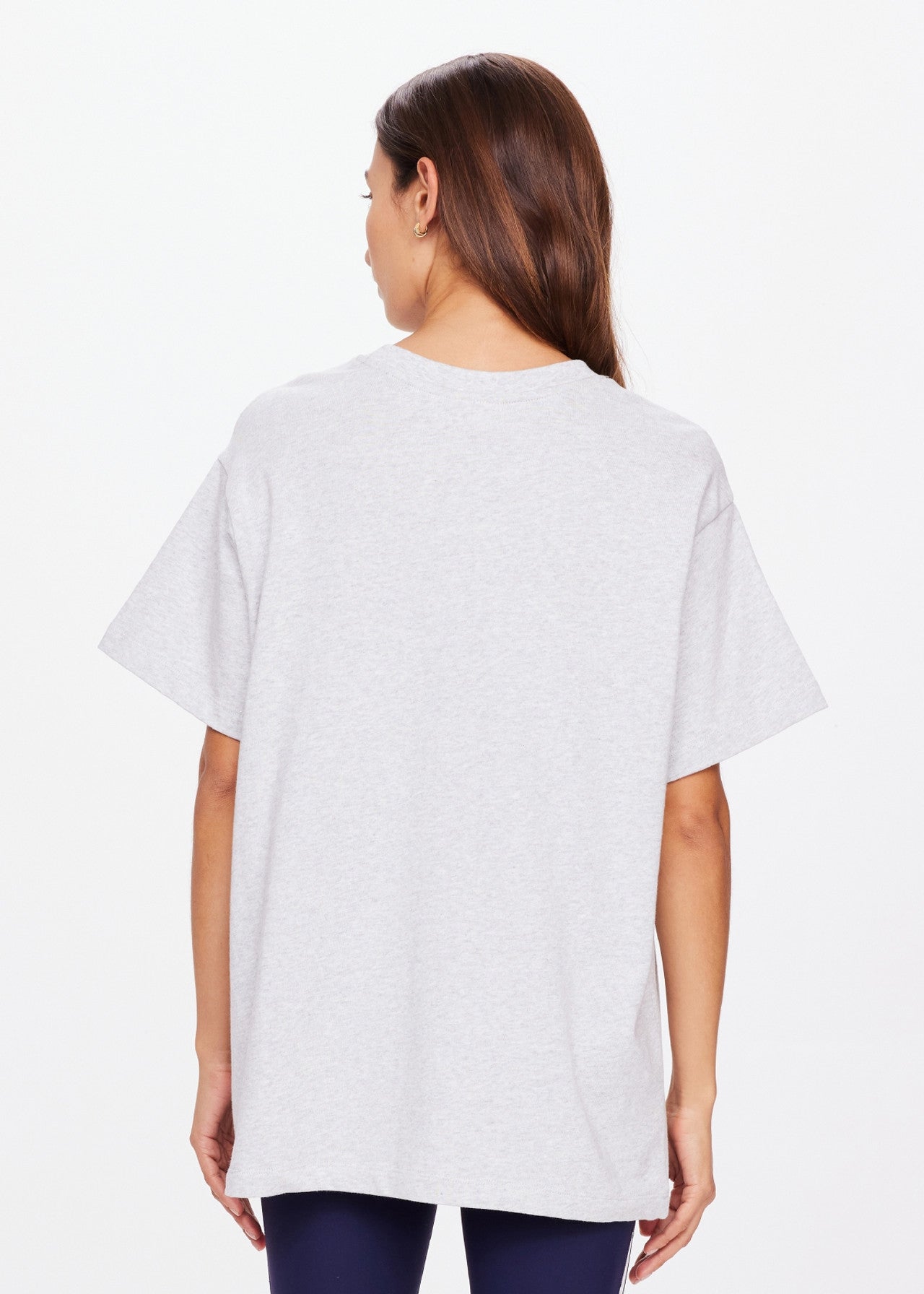 A person with long hair faces away from the camera, wearing The Upside's oversized HARBOR SAMMI TEE made of organic cotton, paired with dark pants against a plain white background.