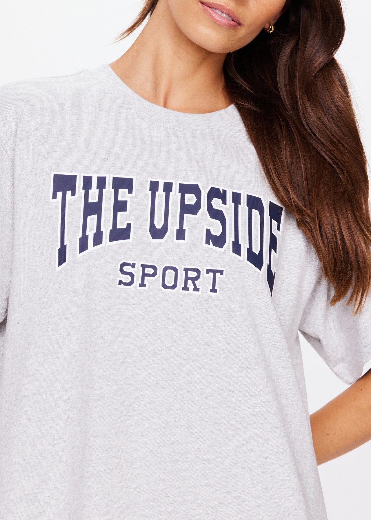 A person is seen in an oversized light gray HARBOR SAMMI TEE from The Upside, crafted from organic cotton and featuring "THE UPSIDE SPORT" in bold, dark blue letters. Their long brown hair drapes over their shoulder as the image focuses on the upper torso and part of the face.