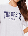 A person is seen in an oversized light gray HARBOR SAMMI TEE from The Upside, crafted from organic cotton and featuring "THE UPSIDE SPORT" in bold, dark blue letters. Their long brown hair drapes over their shoulder as the image focuses on the upper torso and part of the face.