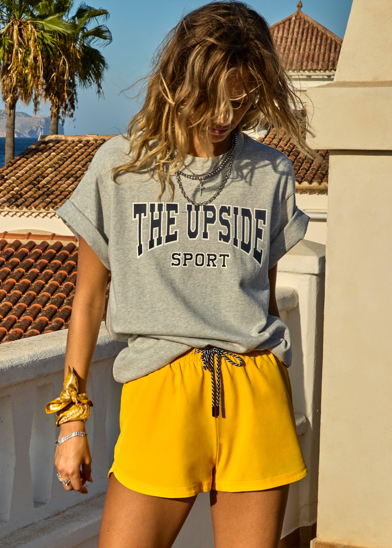 A woman with wavy hair wears a gray "HARBOR SAMMI TEE" from The Upside and yellow shorts. She stands on a terrace amid palm trees and a tiled roof under the clear sky, exuding effortless style in her organic cotton outfit.