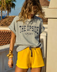 A woman with wavy hair wears a gray "HARBOR SAMMI TEE" from The Upside and yellow shorts. She stands on a terrace amid palm trees and a tiled roof under the clear sky, exuding effortless style in her organic cotton outfit.