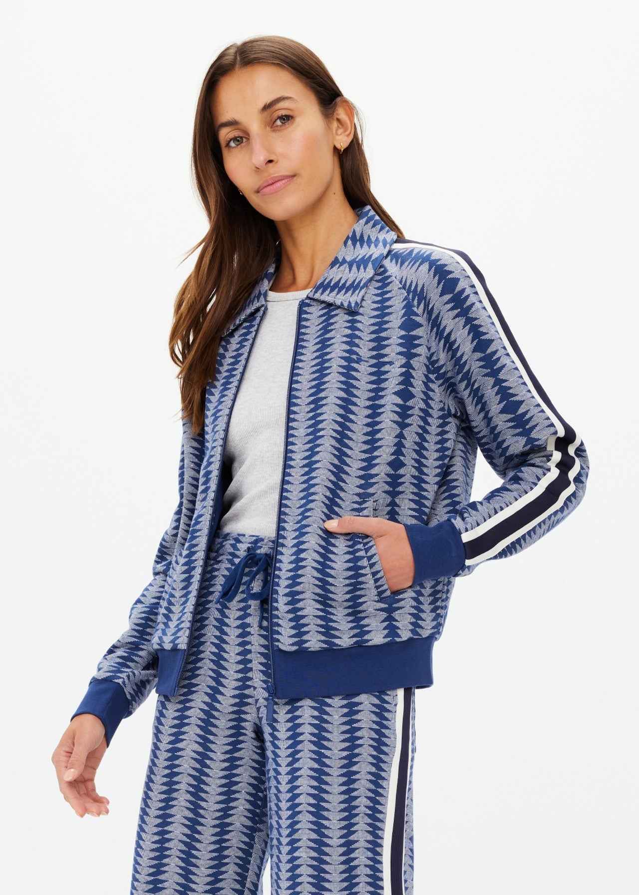 A person with long brown hair is modeling The Upside's JOIE PARI ZIP JACKET, featuring a blue and gray houndstooth pattern, ribbed hems, and knitted stripe sleeves. They are pairing it with matching pants while posing against a plain white background.