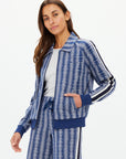 A person with long brown hair is modeling The Upside's JOIE PARI ZIP JACKET, featuring a blue and gray houndstooth pattern, ribbed hems, and knitted stripe sleeves. They are pairing it with matching pants while posing against a plain white background.