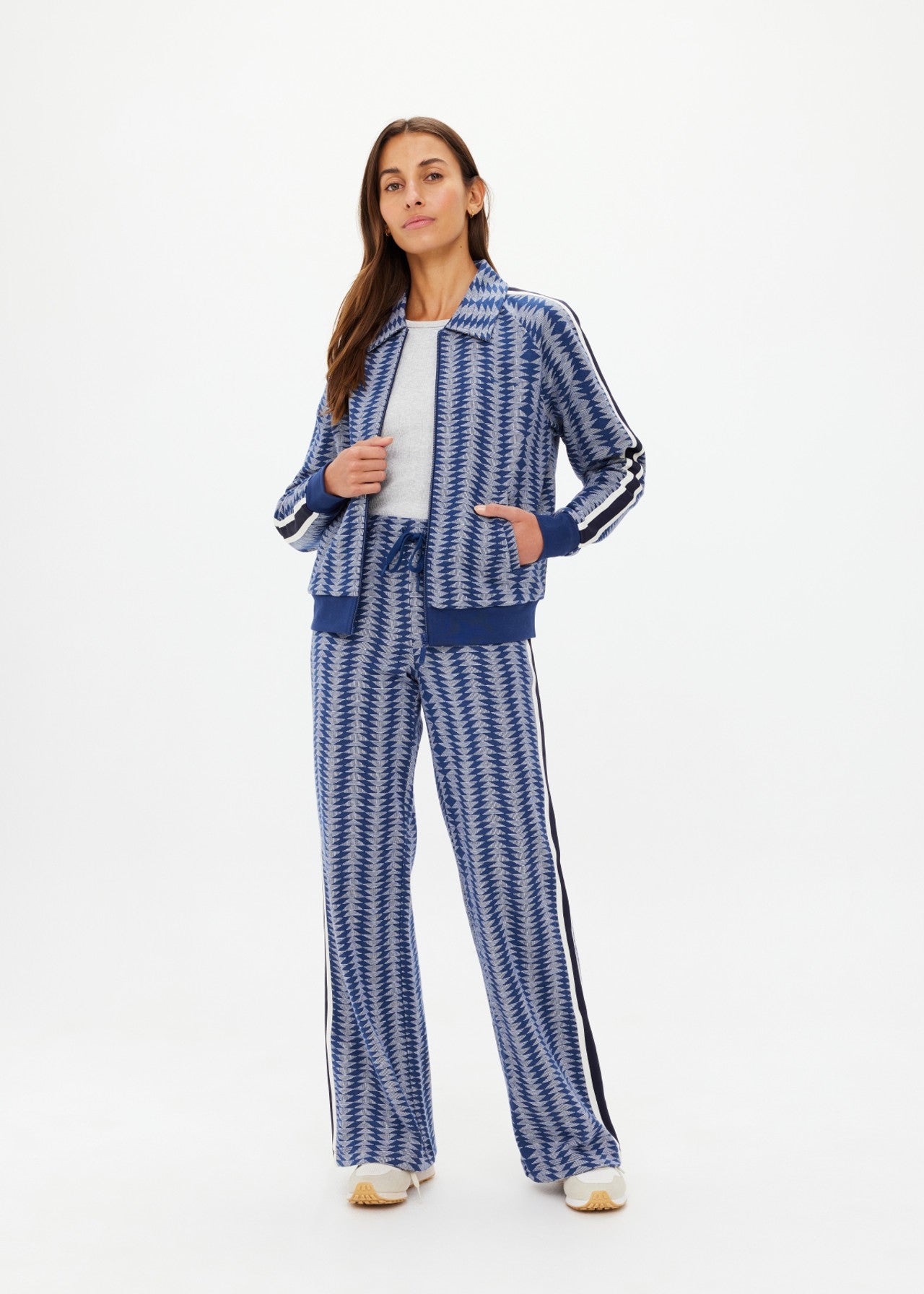 With confidence, a long-haired individual dons The Upside's JOIE PARI ZIP JACKET featuring a blue and white zigzag pattern, knitted stripes, and ribbed hem. Their white top peeks through as they place one hand in their pocket, looking forward with poise.