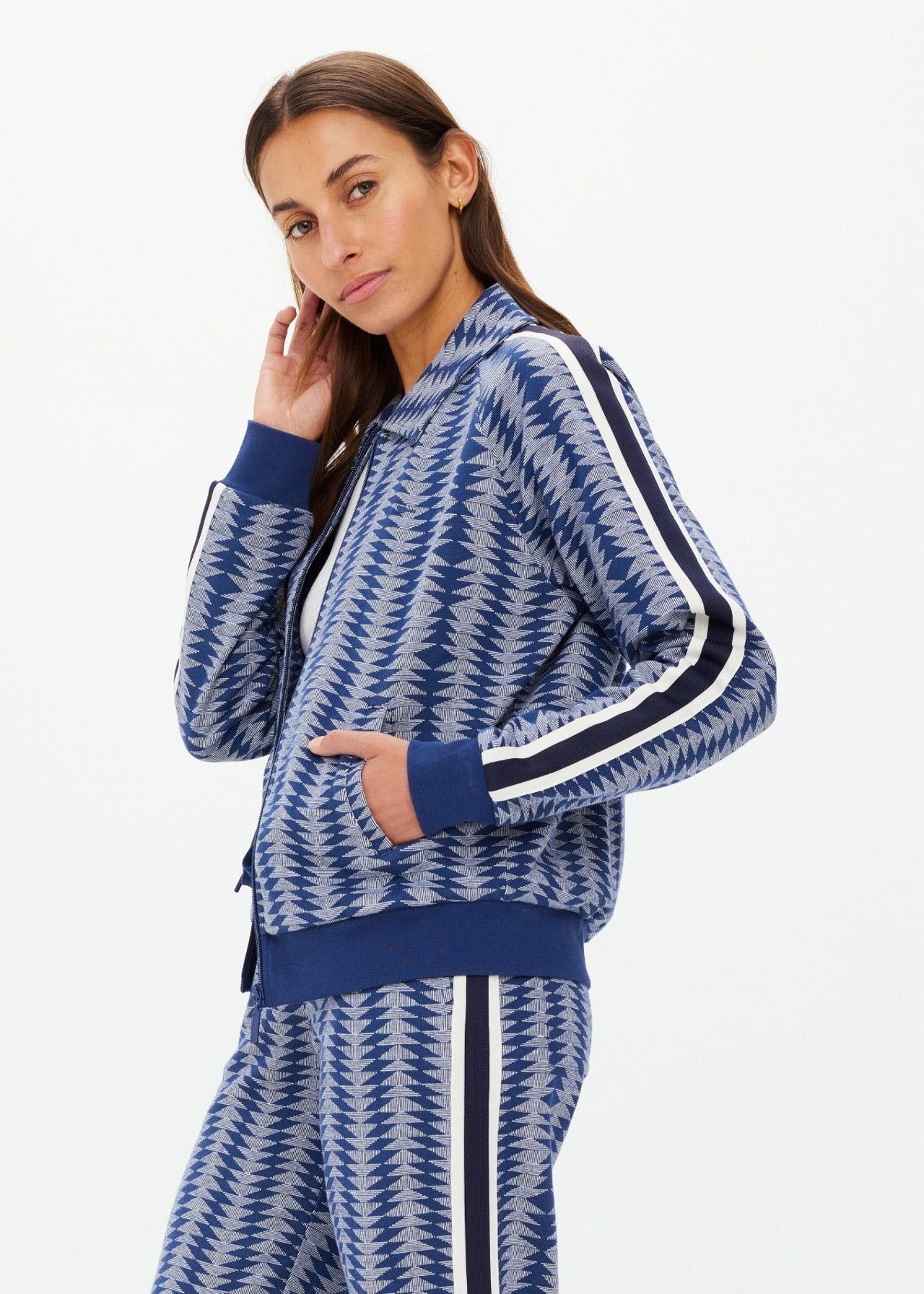 A person with long hair models a JOIE PARI ZIP JACKET by The Upside, a blue patterned tracksuit featuring white and black stripes on the sleeves and pants. They pose sideways against a plain white background, touching their chin.