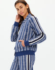 A person with long hair models a JOIE PARI ZIP JACKET by The Upside, a blue patterned tracksuit featuring white and black stripes on the sleeves and pants. They pose sideways against a plain white background, touching their chin.