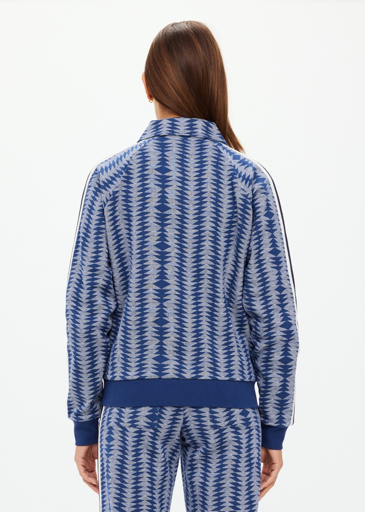 A person with long brown hair, seen from behind, wears The Upside's JOIE PARI ZIP JACKET, a blue and white patterned sports jacket featuring a ribbed hem, knitted sleeve stripes, and a high collar. The background is plain and light-colored.