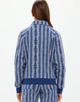 A person with long brown hair, seen from behind, wears The Upside's JOIE PARI ZIP JACKET, a blue and white patterned sports jacket featuring a ribbed hem, knitted sleeve stripes, and a high collar. The background is plain and light-colored.