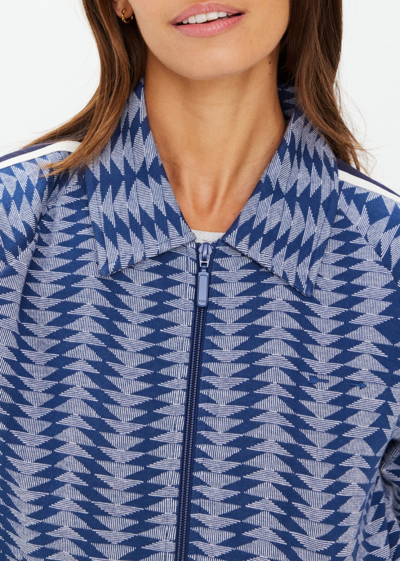 A person sports The Upside's JOIE PARI ZIP JACKET, showcasing light and dark blue chevrons in jacquard knit. Made from organic cotton, it features a wide collar and striped tape on the shoulders. The face is partially visible.