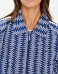 A person sports The Upside's JOIE PARI ZIP JACKET, showcasing light and dark blue chevrons in jacquard knit. Made from organic cotton, it features a wide collar and striped tape on the shoulders. The face is partially visible.