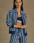 Wearing The Upside's JOIE PARI ZIP JACKET in a striking blue and white Jacquard design, paired with matching pants and sports bra. Their long dark hair complements gold earrings and bracelets, set against a neutral background.