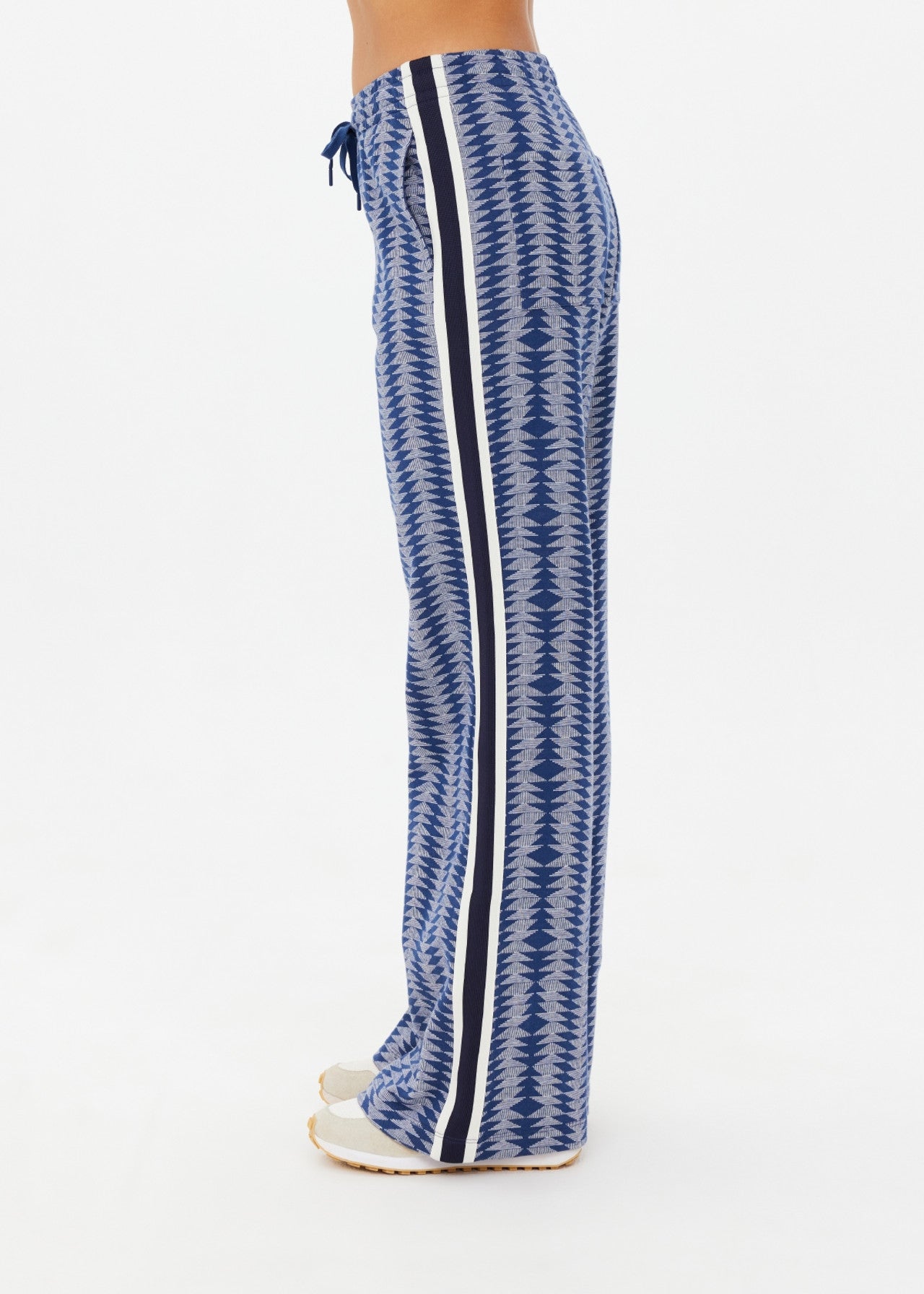 A person is seen from the side wearing The Upside's JOIE JETSET STRAIGHT LEG PANT, crafted from an organic cotton blend with a blue pattern and black and white side stripes. They are paired with light-colored shoes, standing on a light background.