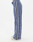 A person is seen from the side wearing The Upside's JOIE JETSET STRAIGHT LEG PANT, crafted from an organic cotton blend with a blue pattern and black and white side stripes. They are paired with light-colored shoes, standing on a light background.