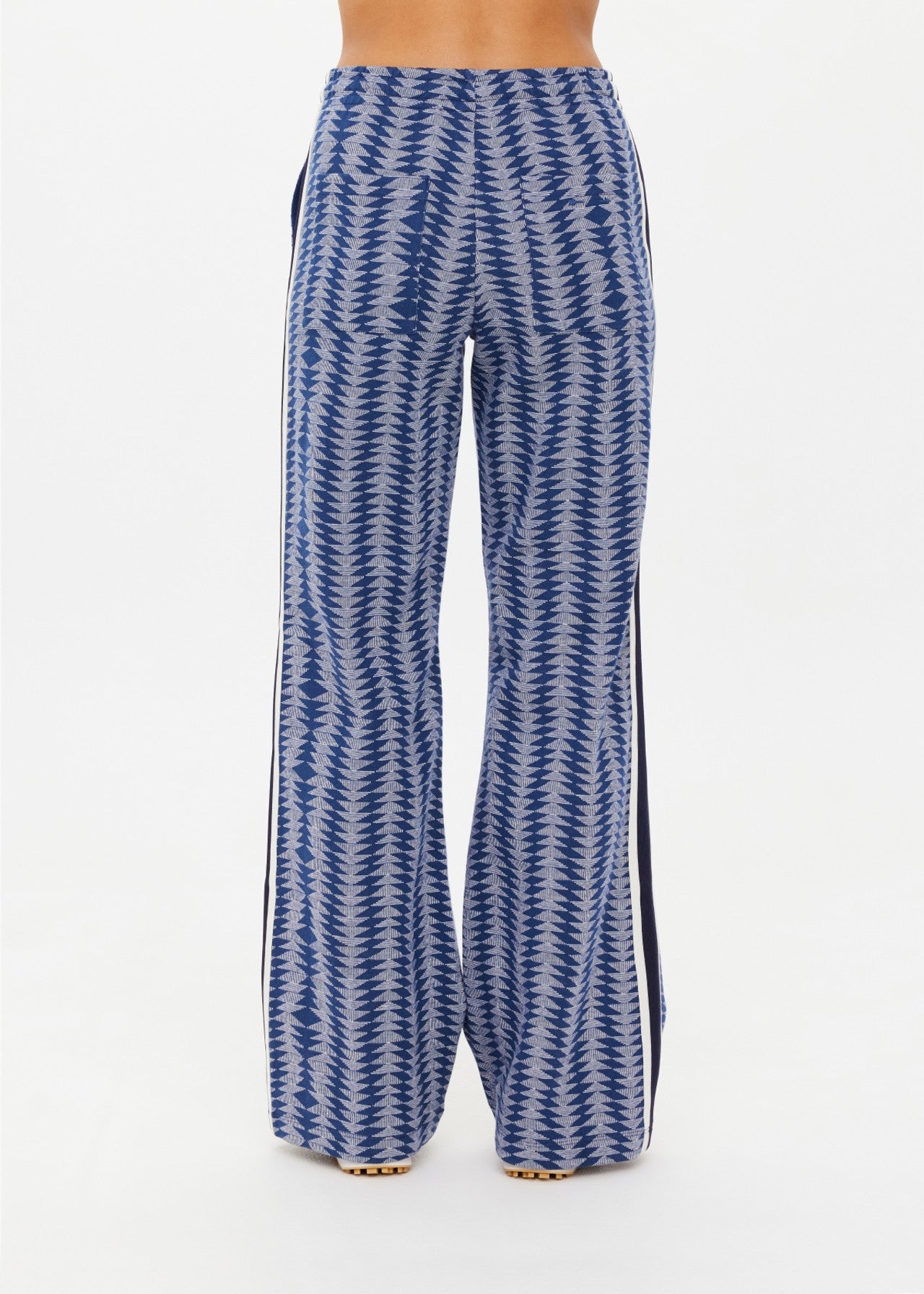 Standing against a plain white background, a person wears The Upside JOIE JETSET STRAIGHT LEG PANT, featuring a blue and white geometric jacquard pattern with sleek black vertical side stripes.