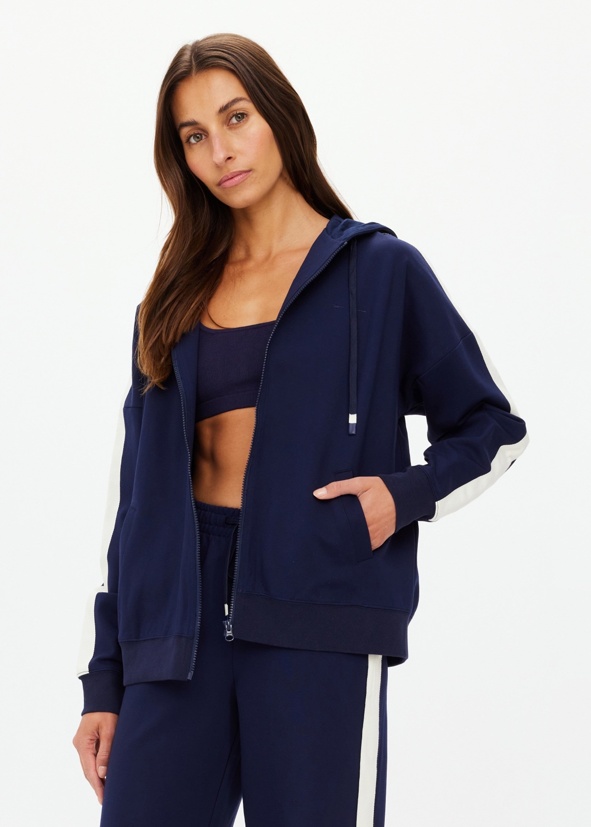 A woman with long brown hair wears The Upside Hamptons Ali Zip Up Hoodie, featuring white sleeve stripes and an embroidered logo, along with a matching sports bra and navy blue pants. She stands against a plain white background, looking at the camera.