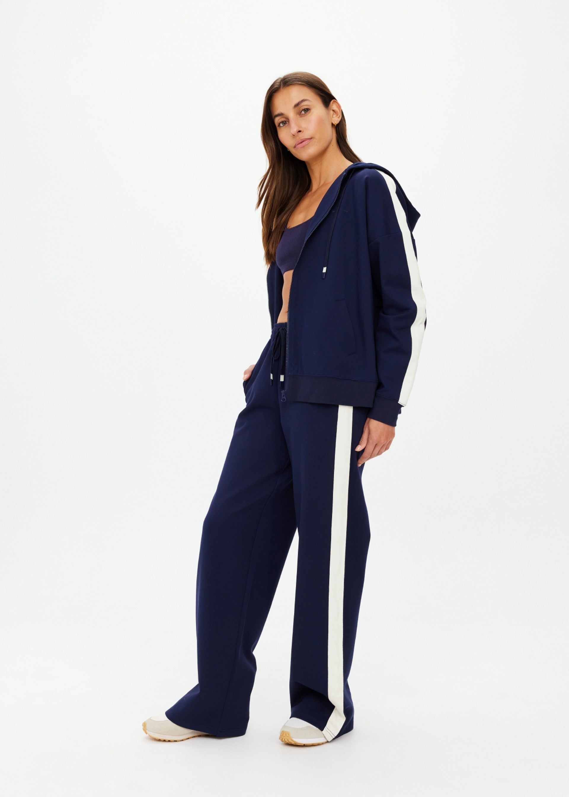 A woman stands in a relaxed pose wearing The Upside's Hamptons Ali Zip Up Hoodie in navy, styled with matching wide-leg pants and a navy sports bra, set against a plain white background.