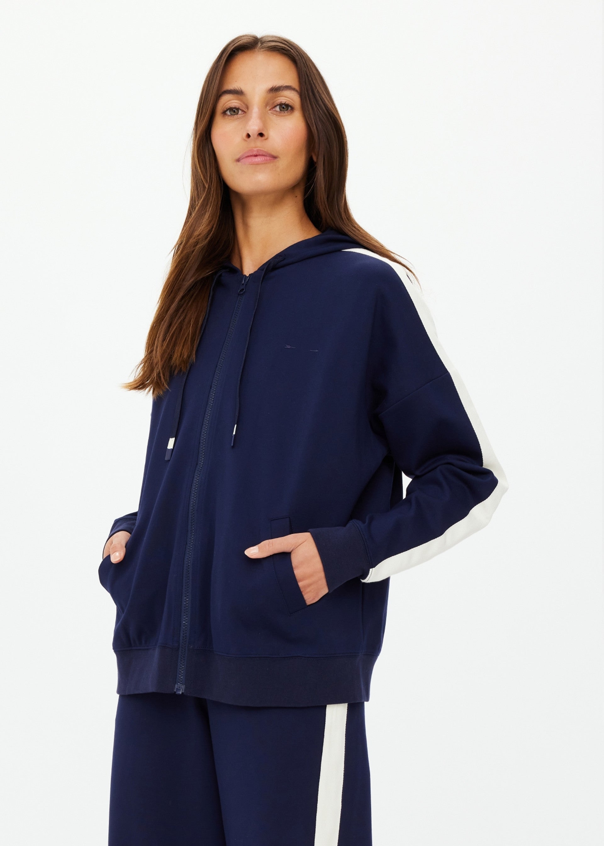 A woman wears The Upside Hamptons Ali Zip Up Hoodie in navy with white sleeve stripes and an embroidered tonal arrow logo, paired with matching navy pants with white side stripes, standing hands in pockets against a plain white background.