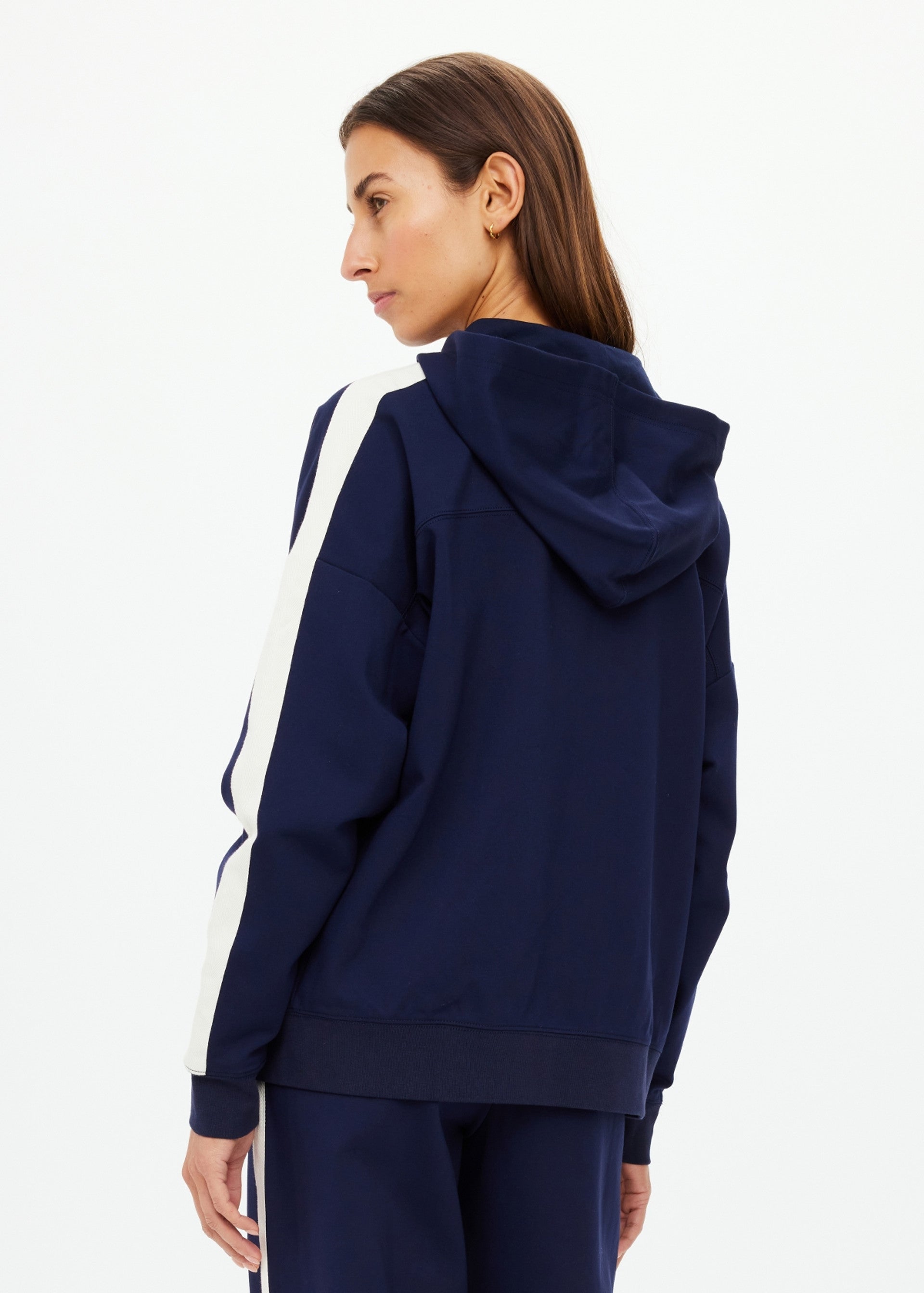 A woman with long brown hair models The Upside Hamptons Ali Zip Up Hoodie in navy blue with white sleeve stripes and a tonal arrow logo, turning her head slightly while standing against a plain white background.