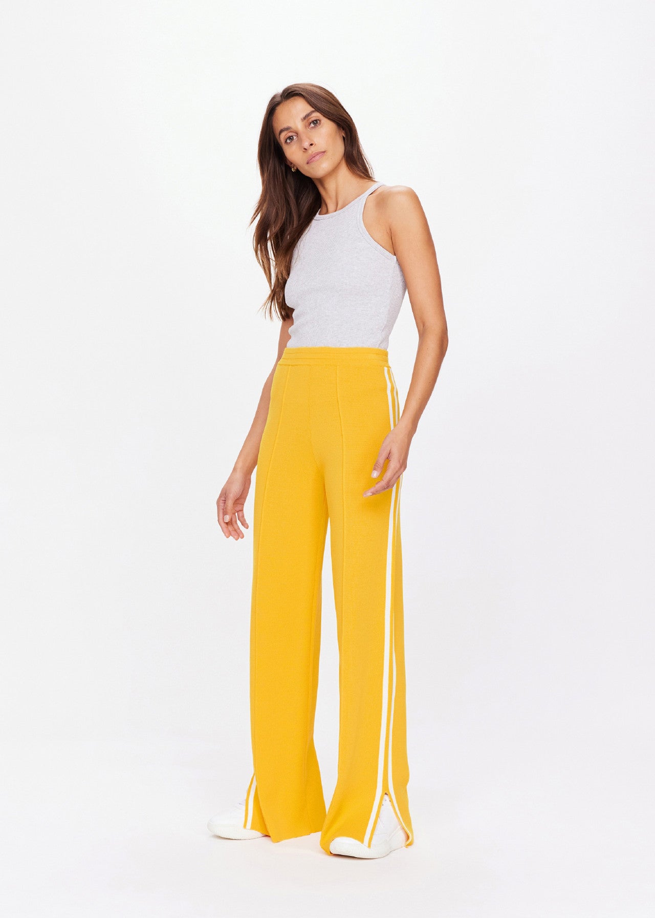 A woman exudes sporty sophistication in a gray sleeveless top and The Upside's Dunes Freya Knit Pant, featuring marigold wide-leg style with white stripes. Her long brown hair cascades down as she poses against a white background, completing the look with crisp white shoes.