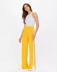 A woman exudes sporty sophistication in a gray sleeveless top and The Upside's Dunes Freya Knit Pant, featuring marigold wide-leg style with white stripes. Her long brown hair cascades down as she poses against a white background, completing the look with crisp white shoes.