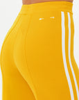 Close-up of a person wearing The Upside's Dunes Freya Knit Pant in vibrant marigold, featuring two white side stripes, a textured fabric, and small white arrow symbols on the waistline, all against a light background.