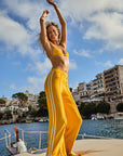 A joyful woman in a vibrant marigold bikini top and The Upside's Dunes Freya Knit Pant dances on the boat deck, set against the serene backdrop of calm water, surrounding boats, and seaside buildings beneath a partly cloudy blue sky.