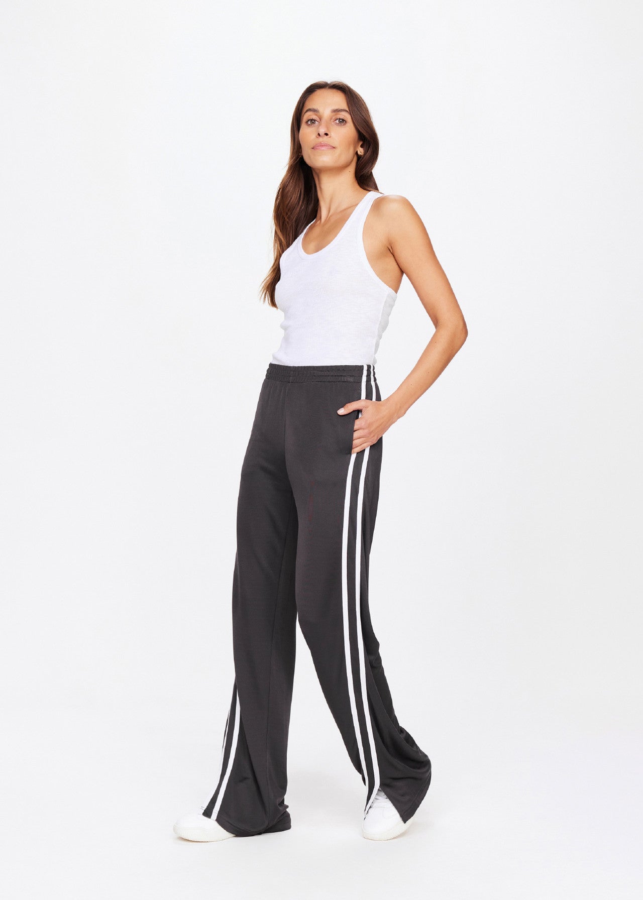 Wearing a white tank top and The Upside's JULIET PANT with a chic low rise, wide legs, and white side stripes, they exude confidence. Long hair complements the style, paired perfectly with crisp white sneakers against a simple backdrop.