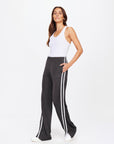 Wearing a white tank top and The Upside's JULIET PANT with a chic low rise, wide legs, and white side stripes, they exude confidence. Long hair complements the style, paired perfectly with crisp white sneakers against a simple backdrop.