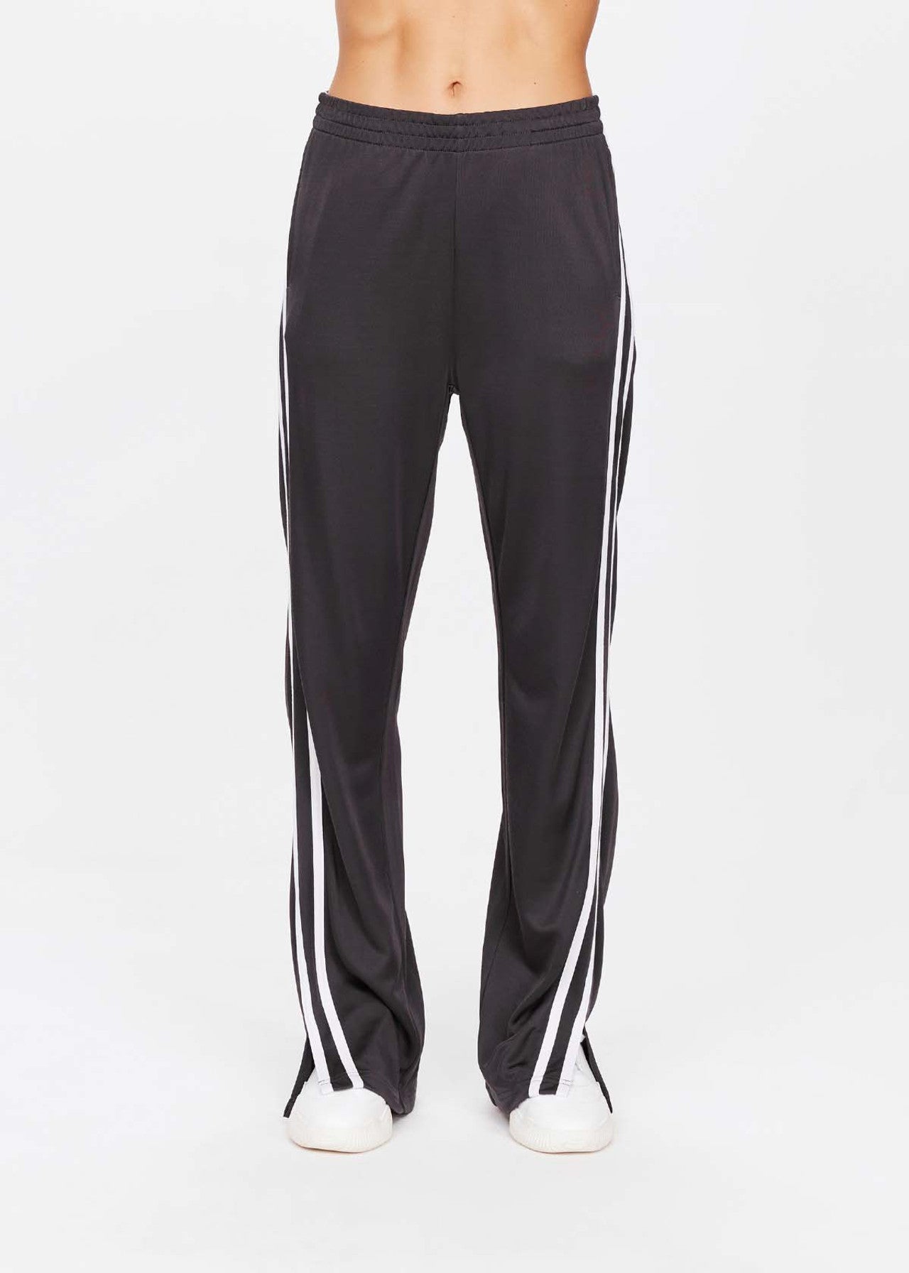 A person wears The Upside's JULIET PANT, sleek black track pants with white side stripes and chic white shoes. Set against a plain white backdrop, these low-rise pants are loose-fitting, expertly cropped at the waist and below the ankles for a modern style.