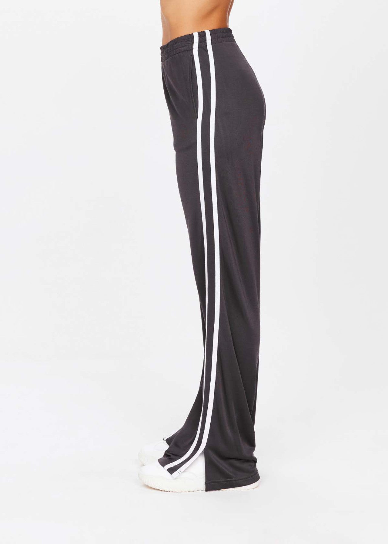 A side view shows someone wearing The Upside's Juliet Pant, black and low-rise with white side stripes, against a white background. Made from slinky fabric, these pants complement sleek white shoes perfectly.