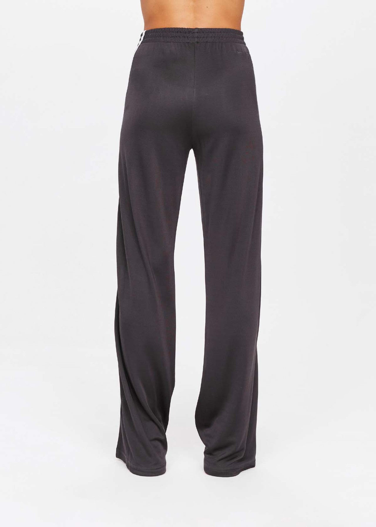 Someone stands against a plain white background wearing The Upside's JULIET PANT, loose-fitting dark gray pants made with slinky fabric. Featuring an elastic waistband and flowy legs, these pants exude relaxed elegance. The upper body is not visible.