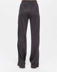 Someone stands against a plain white background wearing The Upside's JULIET PANT, loose-fitting dark gray pants made with slinky fabric. Featuring an elastic waistband and flowy legs, these pants exude relaxed elegance. The upper body is not visible.