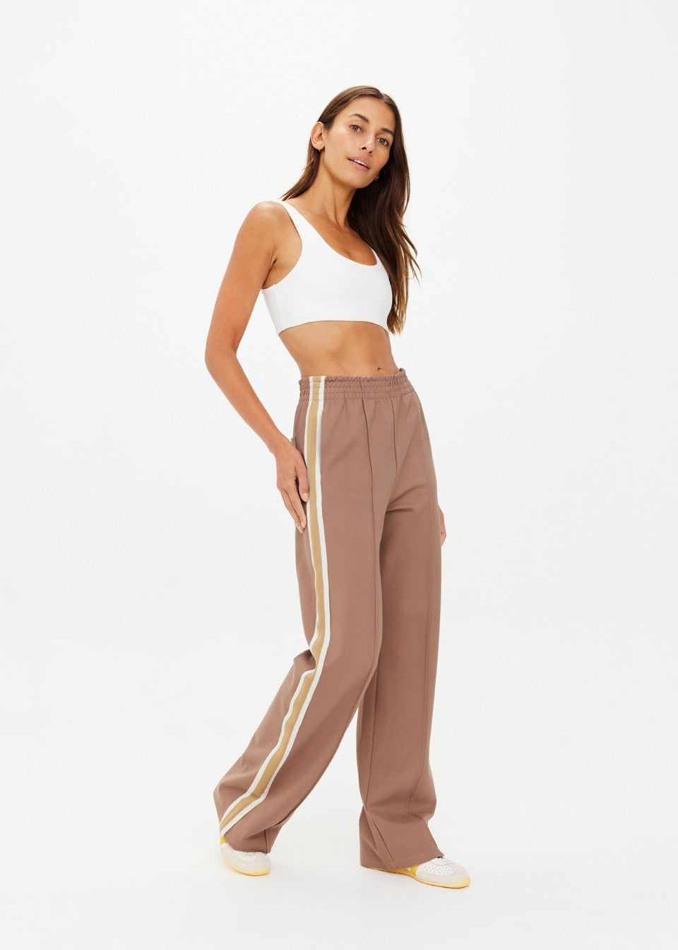 A person confidently poses in The Upside's MONTE PANTS, high-waisted with white side stripes. Paired with a white crop top and stylish white shoes with yellow details, their long hair cascades against a plain background, achieving an effortlessly chic look.