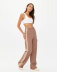 A person confidently poses in The Upside's MONTE PANTS, high-waisted with white side stripes. Paired with a white crop top and stylish white shoes with yellow details, their long hair cascades against a plain background, achieving an effortlessly chic look.